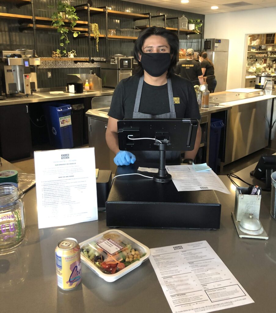 Photo of a barista with face mask looking at the camera, at the register in a cafe.