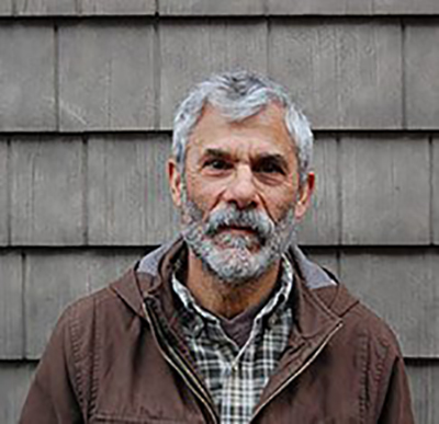 Headshot of an older man with a beard, in a jacket, standing in front of a wood shingled wall.