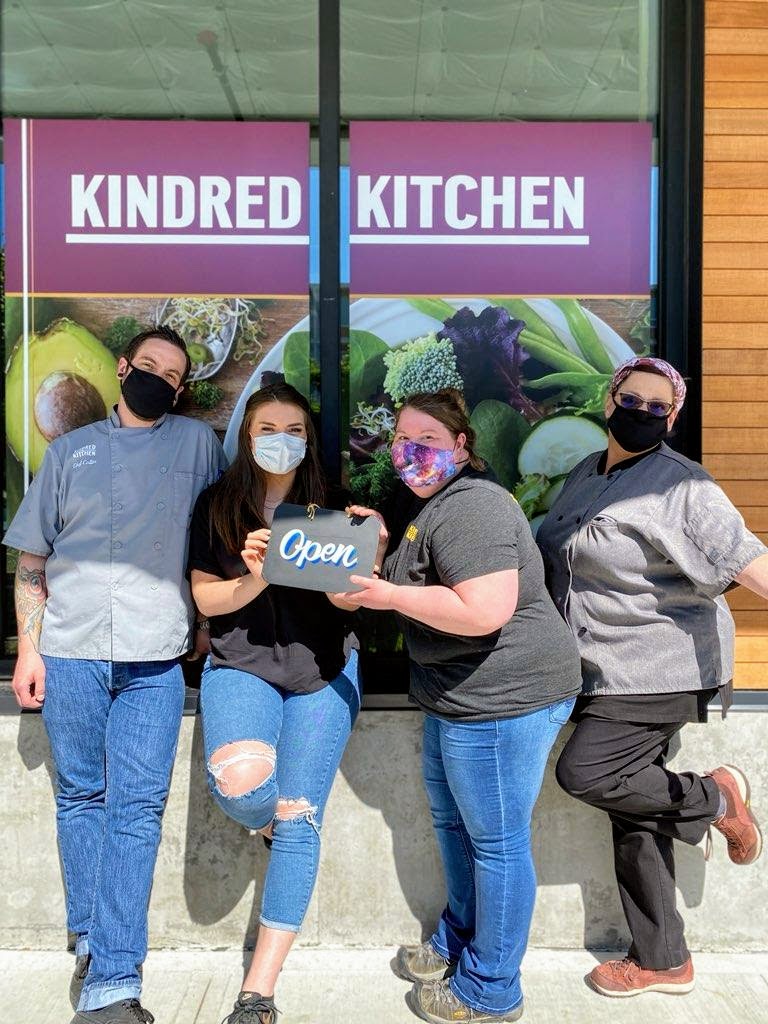 Photo of four people in face masks, holding a small open sign in front of Kindred Kitchen.