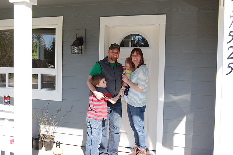 Photo of a family with two small children, in front of a doorway leading into a home.
