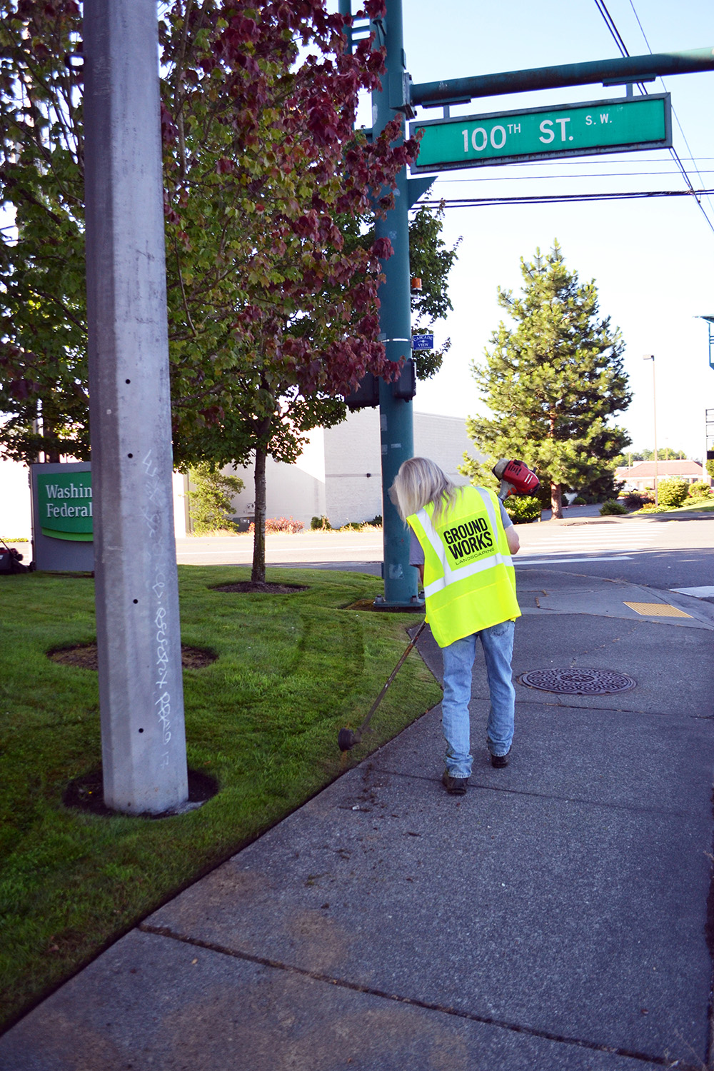 Photo of a man with long hair, weedwhacking a commercial greenway near the corner of an intersection.