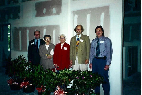 Older photo of five people in formal wear, standing in front of an unpainted wall and behind small shrubs, looking at the camera.