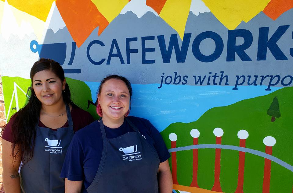 Photo of two women in aprons, standing in front of a CafeWorks mural.