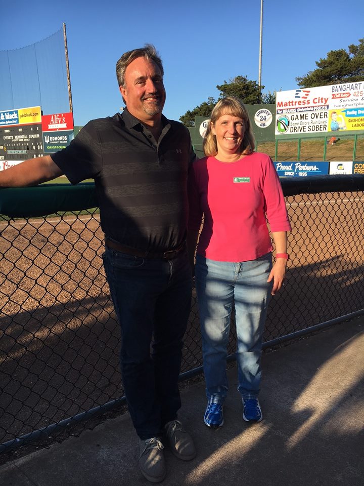 Photo of a man and woman in casual attire, leaning against a chain-link fence in front of a baseball field.