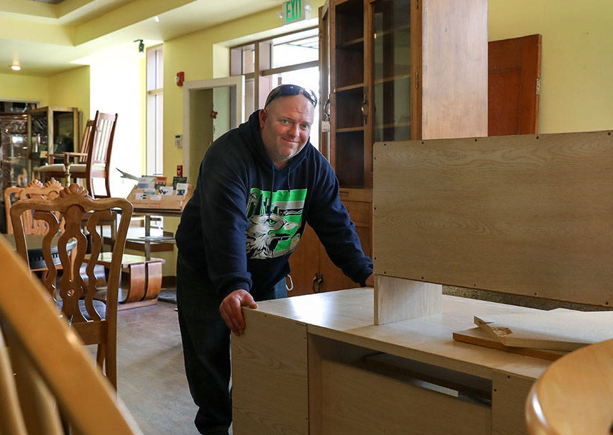 Photo of a man leaning over a desk being refurbished, smiling at the camera.