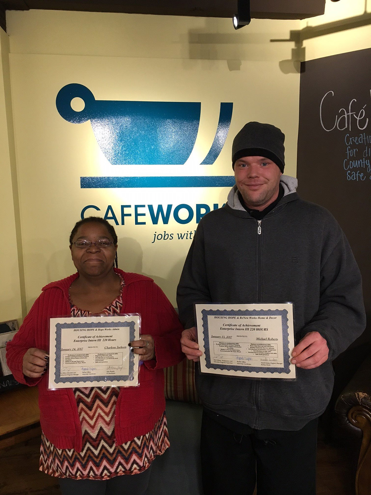 Photo of a man and a woman smiling at the camera, holding graduation certificates, with CafeWorks signage behind them.