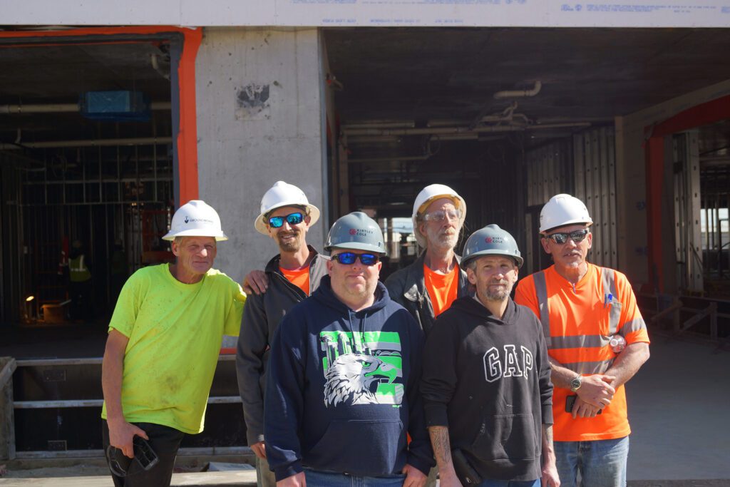 Photo of six men in hardhats, standing in front of a building under construction.