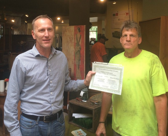 Photo of two men indoors, one holding up a certificate for the other.