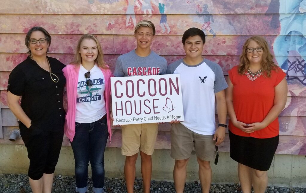 Photo of two women with three teens standing between them, the middle one holding a sign for Cocoon House.