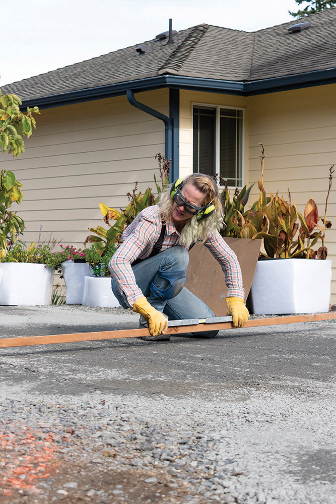 Photo of a woman wearing protective eye and ear gear, kneeling over with a plank of wood, in a driveway of a house.