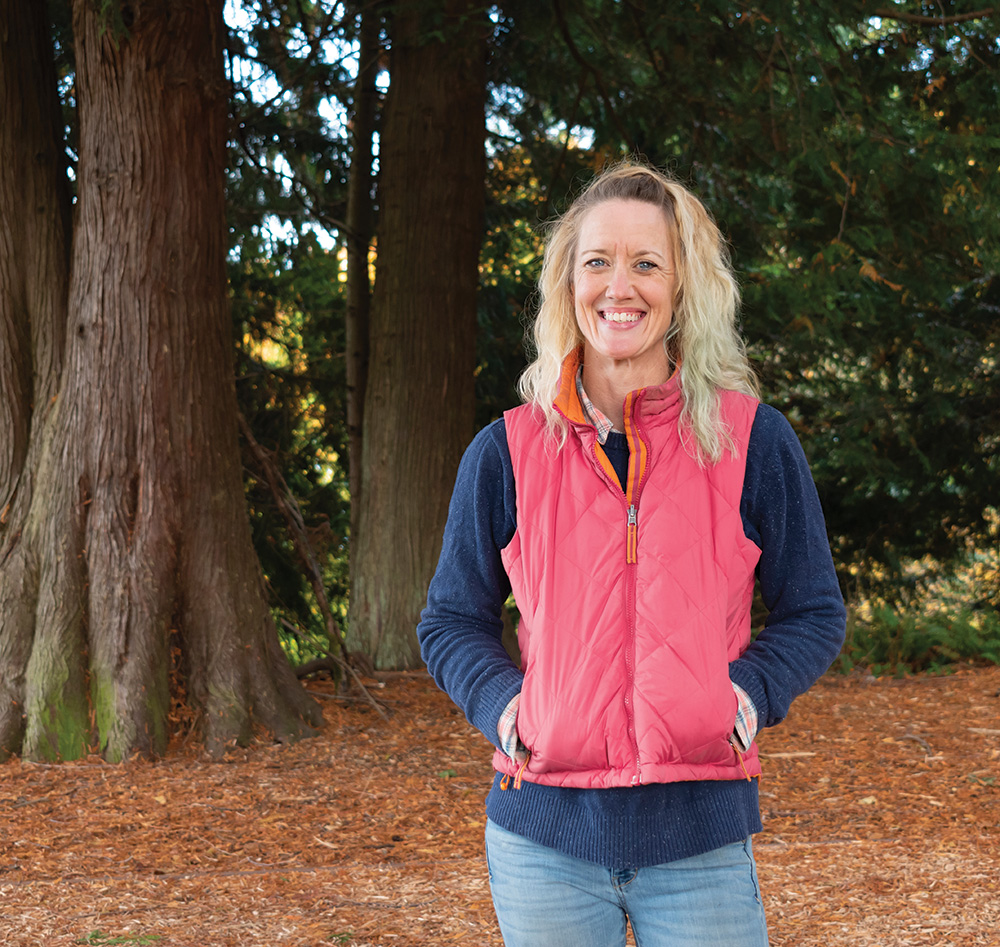 Photo of a woman in a red vest, with hands in pockets, smiling at the camera in a forest environment.