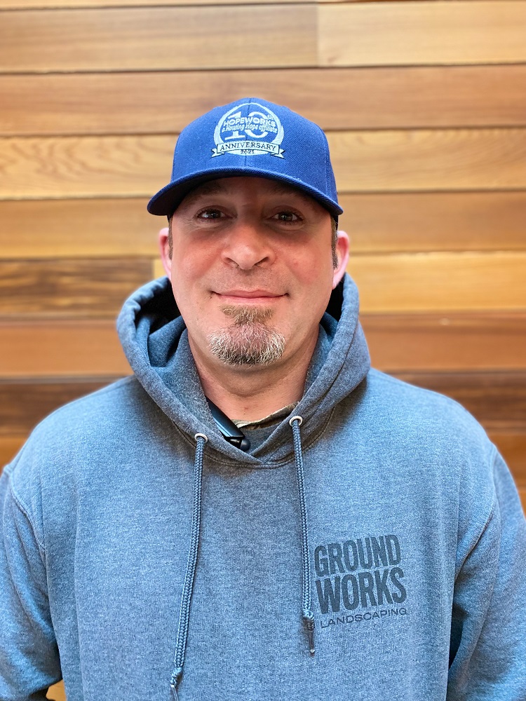 Photo of a man with a goatee in a baseball cap and hoodie, standing in front of a wood wall, looking at the camera.