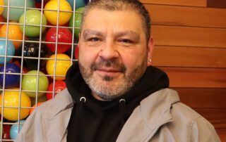 Photo of a man in a hoodie and coat, standing in front of a wood wall and colorful ball sculpture.