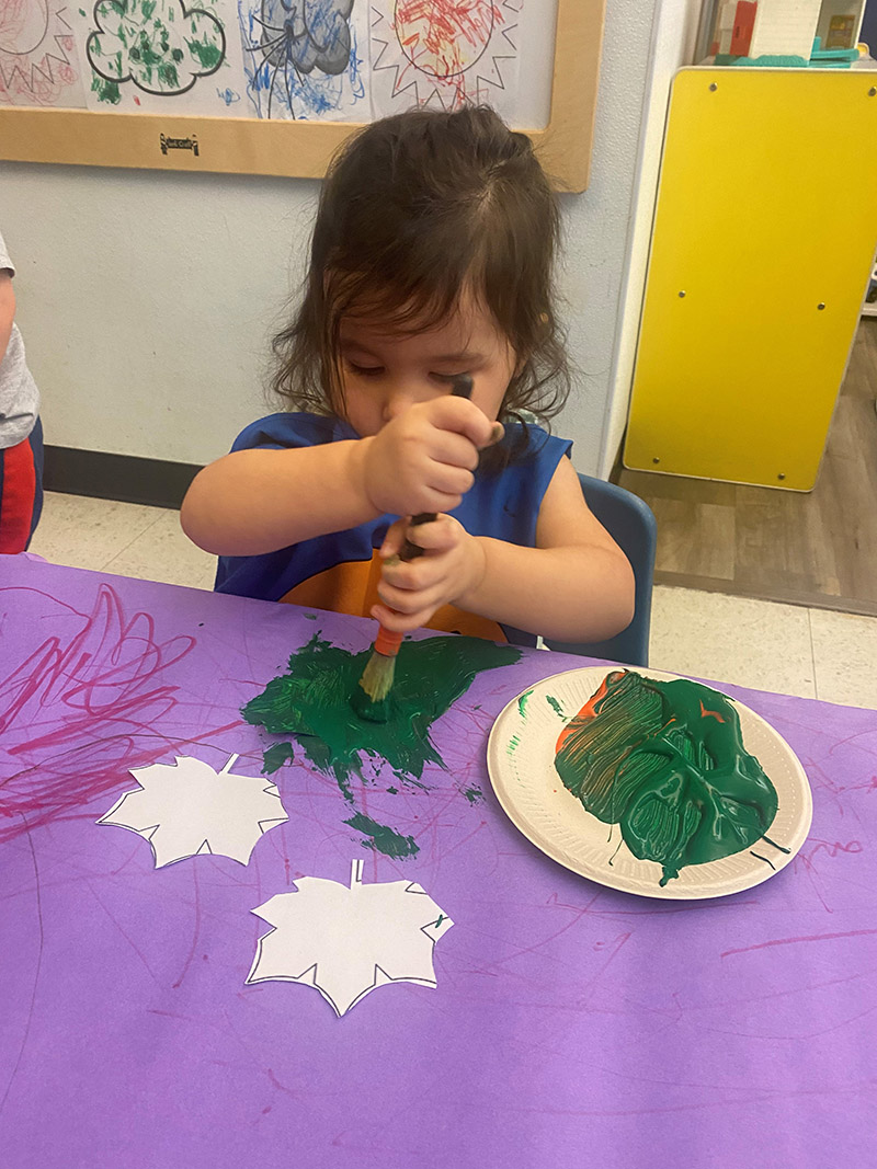 Photo of a toddler playing with a paintbrush and green paint on a paper tablecloth.