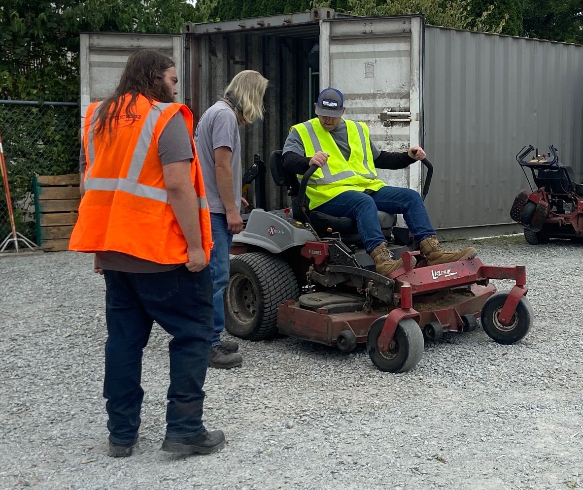 Photo of a man sitting on an industrial mower in a gravel lot, with two other men standing around him.
