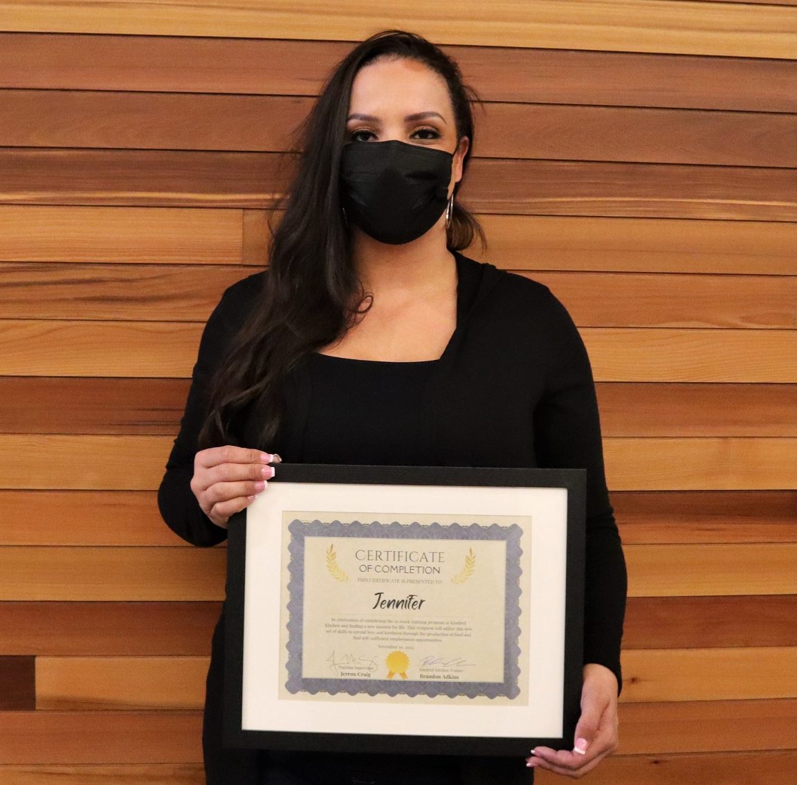 Photo of a woman in a black face mask looking at the camera, holding a graduation certificate.