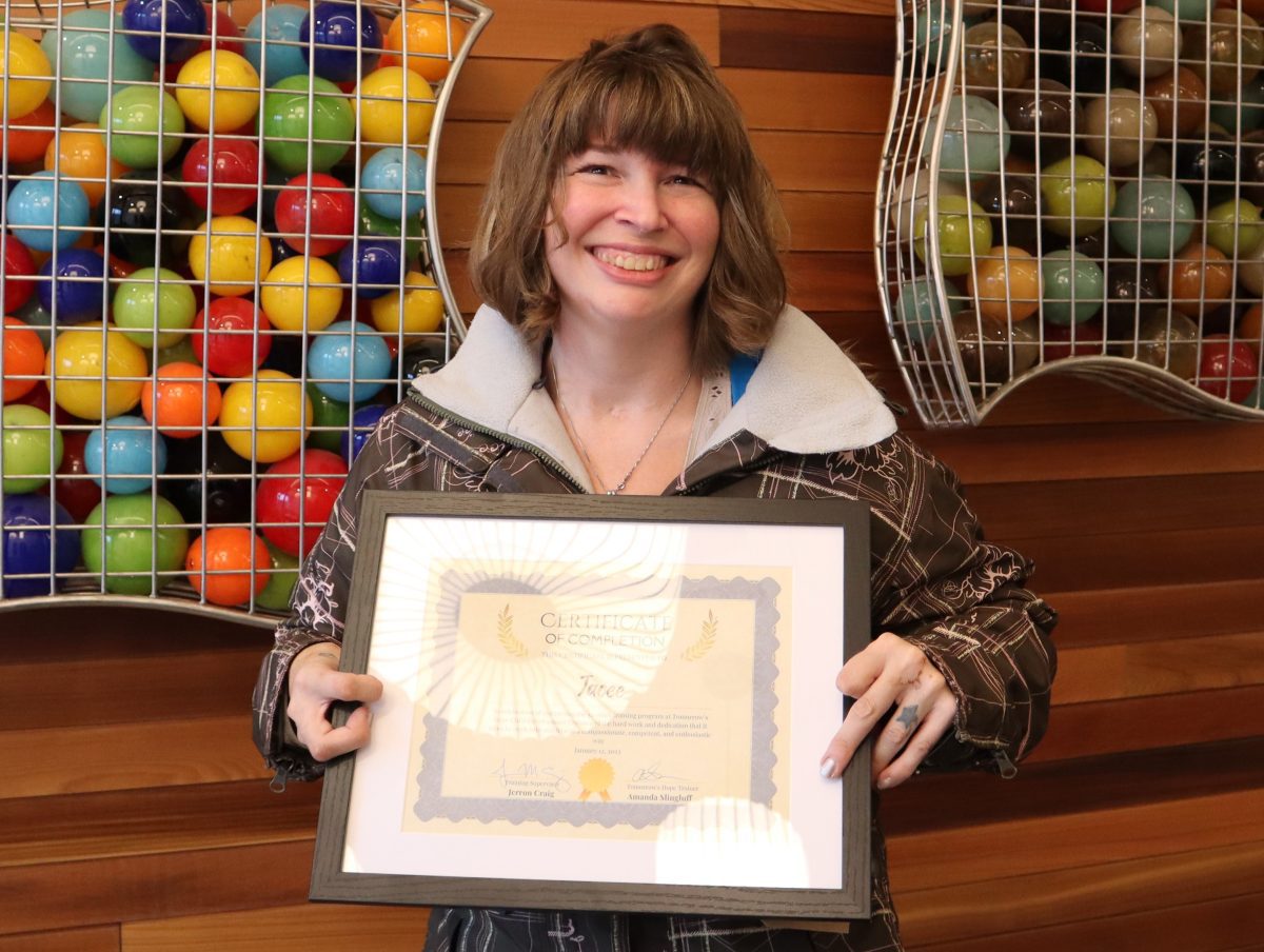 Photo of a smiling woman with a graduation certificate standing in front of a wood wall with colorful balls in cages.