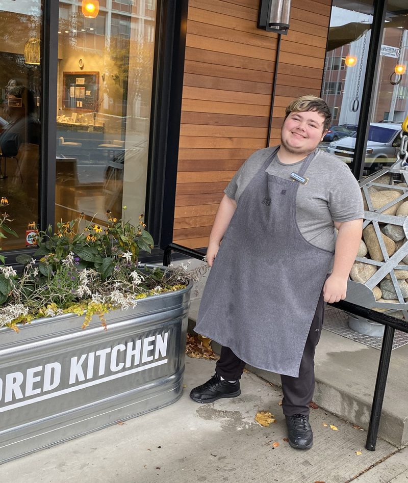 Photo of a young man in an apron standing outside a cafe next to a planter box.