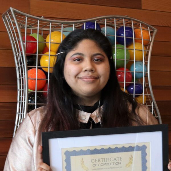 Photo of a woman holding a graduation certificate in front of a wood wall with an artful cage holding colored balls.