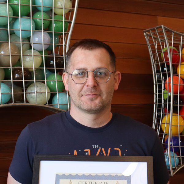 Photo of a man with glasses holding a graduation certificate in front of a wood wall with an art installation with colorful balls.