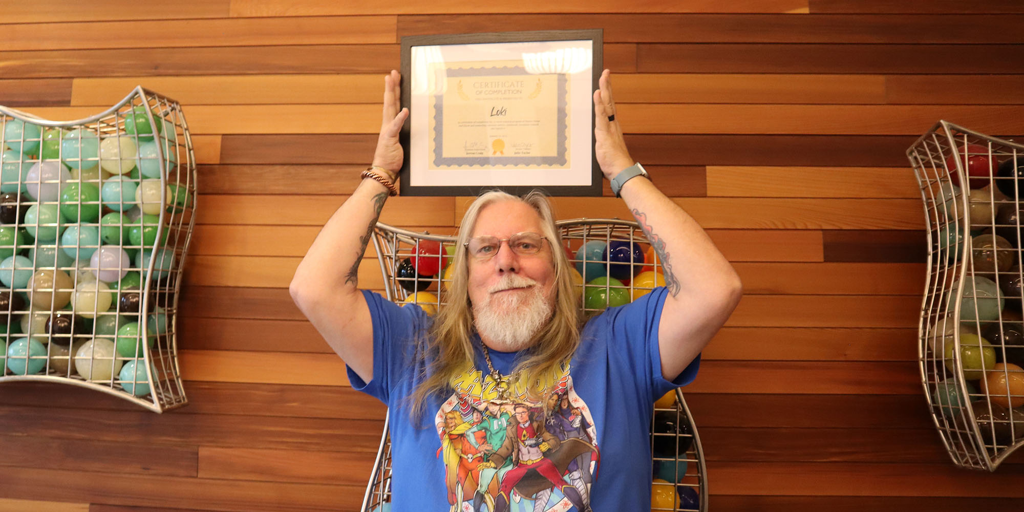 Photo of a man with long hair and a beard holding a graduation certificate above his head, in front of a wood wall.