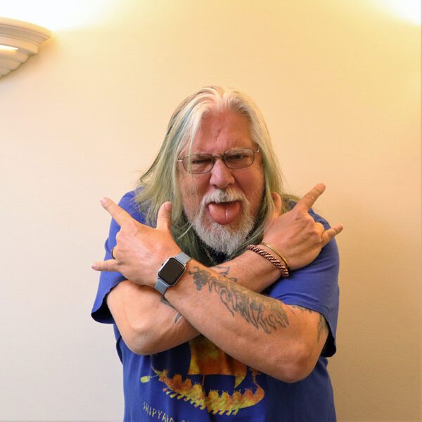 Photo of a man with long hair and beard crossing his arms and sticking out his tongue at the camera.